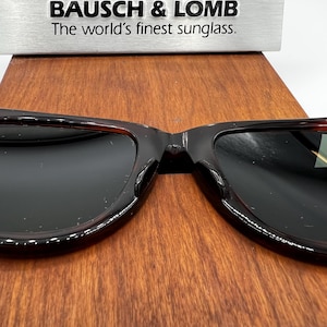 Puede incluir: Gafas de sol con lentes oscuras y montura carey se muestran sobre una superficie de madera. El texto "BAUSCH & LOMB The world's finest sunglass." est&aacute; encima. Las gafas de sol son de estilo cl&aacute;sico.