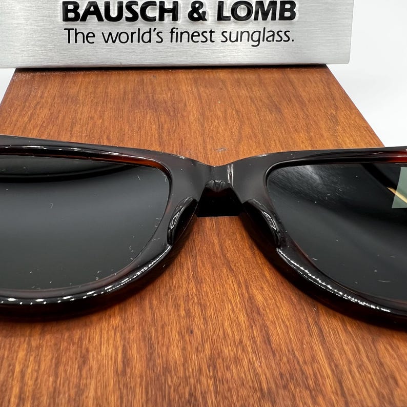 Puede incluir: Gafas de sol con lentes oscuras y montura carey se muestran sobre una superficie de madera. El texto "BAUSCH & LOMB The world's finest sunglass." est&aacute; encima. Las gafas de sol son de estilo cl&aacute;sico.