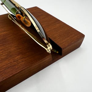 May include: A pair of sunglasses with a black frame and gold-colored accents rests on a wooden block. The lenses are a dark green color. The sunglasses are angled, showcasing their design and the wooden surface.