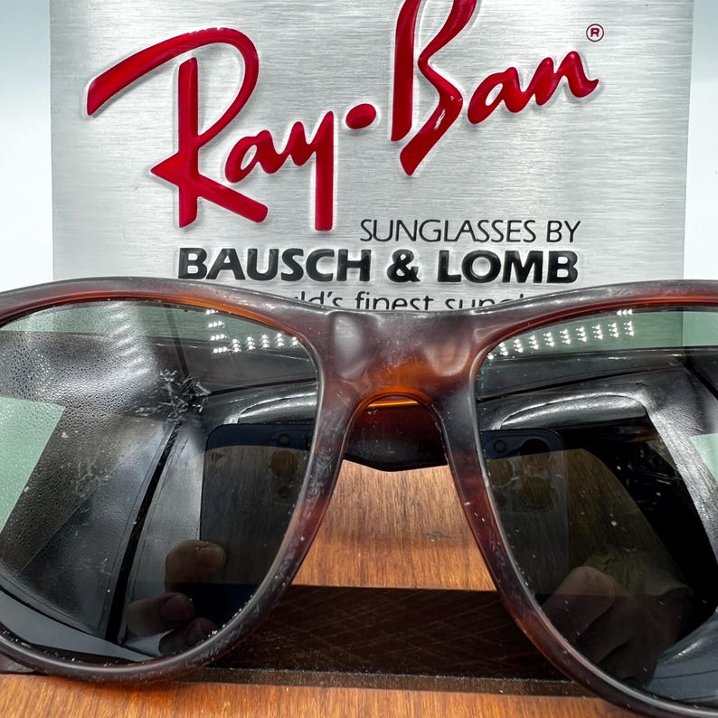 May include: Close-up of Ray-Ban sunglasses with a tortoiseshell frame. The lenses are dark, and the Ray-Ban logo is visible in red on a silver background. The text "SUNGLASSES BY BAUSCH & LOMB" is also displayed.