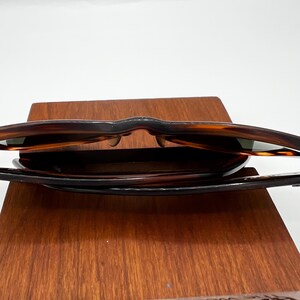 May include: Sunglasses with a tortoiseshell pattern, resting on a wooden display. The sunglasses have dark lenses and a classic frame design. The wooden display has a rich, warm tone.