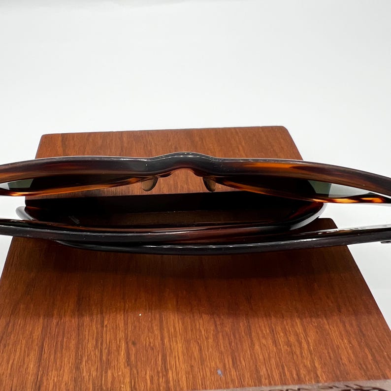 May include: Sunglasses with a tortoiseshell pattern, resting on a wooden display. The sunglasses have dark lenses and a classic frame design. The wooden display has a rich, warm tone.