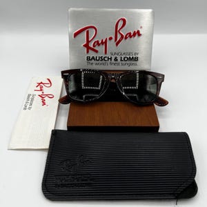 Puede incluir: Gafas de sol Ray-Ban Wayfarer con montura carey oscura, sobre un soporte de madera. Incluye una funda negra, un pa&ntilde;o de limpieza y una pantalla plateada con el logotipo de Ray-Ban y "SUNGLASSES BY BAUSCH & LOMB".