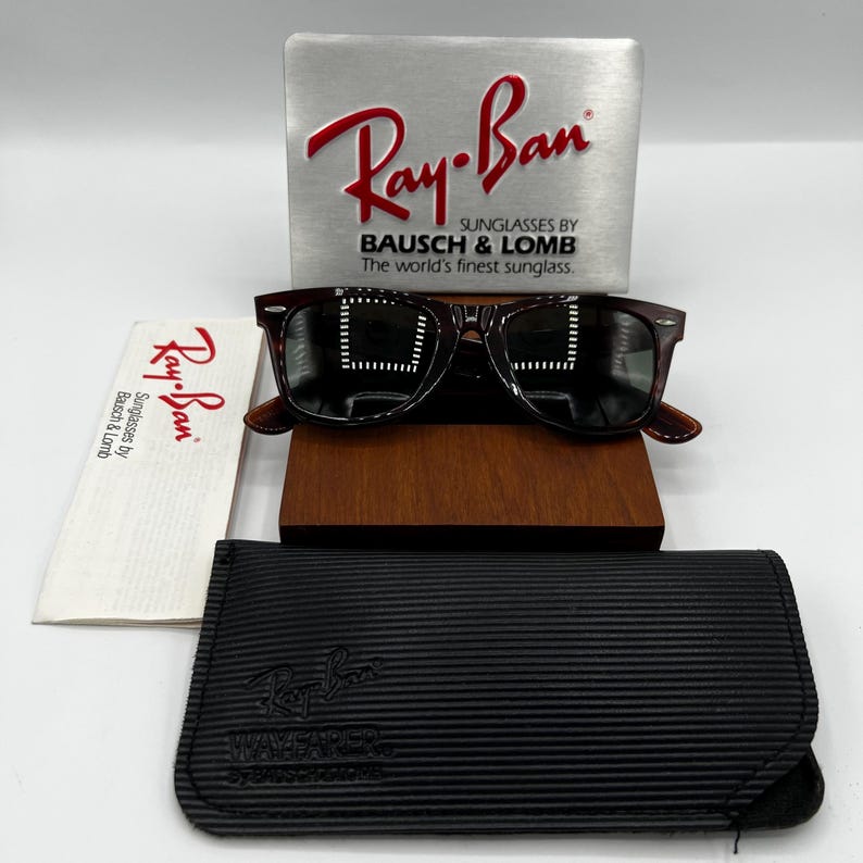 Puede incluir: Gafas de sol Ray-Ban Wayfarer con montura carey oscura, sobre un soporte de madera. Incluye una funda negra, un pa&ntilde;o de limpieza y una pantalla plateada con el logotipo de Ray-Ban y "SUNGLASSES BY BAUSCH & LOMB".