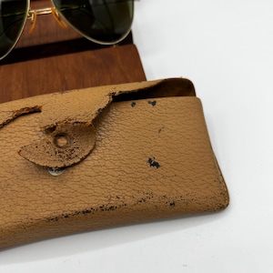 May include: Aviator sunglasses with a gold frame and green lenses rest on a wooden stand. A worn, tan leather case with a snap closure is in the foreground. The case shows signs of wear and tear.