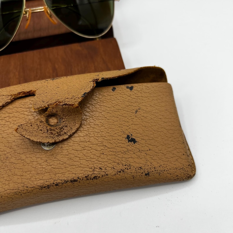 May include: Aviator sunglasses with a gold frame and green lenses rest on a wooden stand. A worn, tan leather case with a snap closure is in the foreground. The case shows signs of wear and tear.