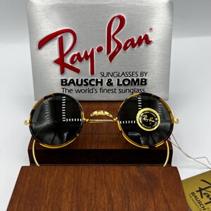 K&ouml;nnte beinhalten: Runde Ray-Ban Sonnenbrille mit Schildpattmuster und goldfarbenen Rahmen. Die Sonnenbrille wird auf einem Holzst&auml;nder pr&auml;sentiert, mit einem silbernen Schild dahinter, auf dem "Ray-Ban" in roter Schrift steht. Das Schild enth&auml;lt auch den Text "SUNGLASSES BY BAUSCH & LOMB".