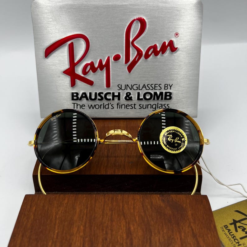K&ouml;nnte beinhalten: Runde Ray-Ban Sonnenbrille mit Schildpattmuster und goldfarbenen Rahmen. Die Sonnenbrille wird auf einem Holzst&auml;nder pr&auml;sentiert, mit einem silbernen Schild dahinter, auf dem "Ray-Ban" in roter Schrift steht. Das Schild enth&auml;lt auch den Text "SUNGLASSES BY BAUSCH & LOMB".