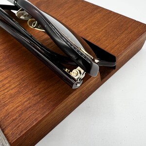 May include: Close-up of black eyeglasses with a dark brown wooden tray. The eyeglasses have a sleek, modern design with a dark frame and gold accents. The tray has a smooth, polished finish.