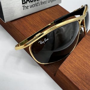 Puede incluir: Gafas de sol Ray-Ban con montura dorada y lentes oscuras sobre una superficie de madera. Las gafas tienen un dise&ntilde;o elegante con puente y varillas negros. El logotipo de Ray-Ban es visible en la lente. El texto "The world's finest sunglasses" es visible en el fondo.