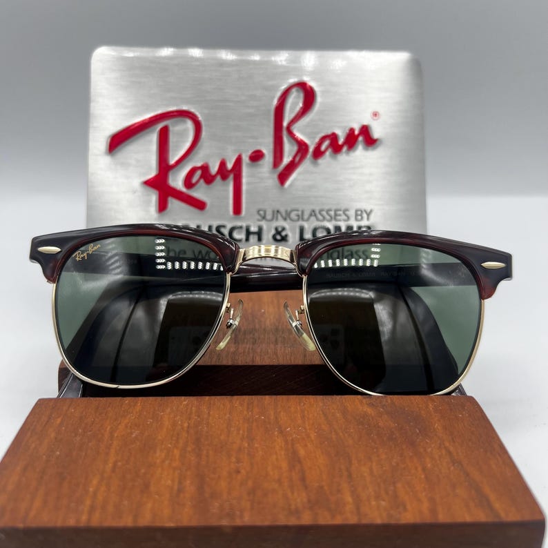 May include: Sunglasses with a dark burgundy frame and gold-colored metal accents. The lenses are dark green. The Ray-Ban logo is visible on the top left of the lens. The sunglasses are displayed on a wooden stand, with the Ray-Ban logo in the background.