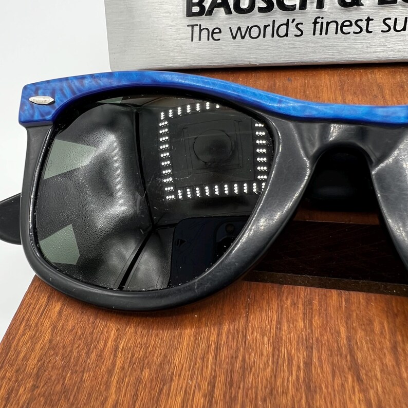 Op de afbeelding: Zwarte zonnebril met een blauw gemarmerd bovenframe. De lenzen zijn donker en reflecteren een heldere lichtbron. De zonnebril ligt op een houten oppervlak.