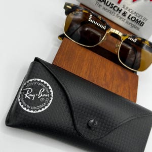 May include: Ray-Ban sunglasses with a tortoiseshell frame and gold-colored accents, resting on a wooden stand. A black, textured case with the Ray-Ban logo is in the foreground. The text "100% UV protection" is visible on the case.
