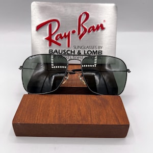 K&ouml;nnte beinhalten: Schwarze Ray-Ban Sonnenbrille mit dunkelgr&uuml;nen Gl&auml;sern, pr&auml;sentiert auf einem Holzst&auml;nder. Das Ray-Ban-Logo ist auf dem oberen linken Glas sichtbar. Die Sonnenbrille befindet sich vor einem silbernen Hintergrund mit dem Ray-Ban-Logo und dem Text "SUNGLASSES BY BAUSCH & LOMB".