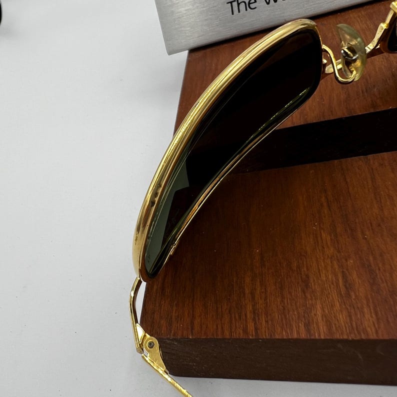 Puede incluir: Gafas de sol con montura dorada y lentes oscuras. Las gafas de sol descansan sobre una superficie de madera oscura. La montura tiene un dise&ntilde;o elegante y moderno. Las lentes son de color oscuro.