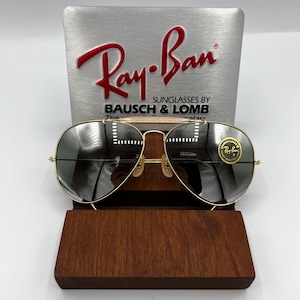May include: Gold-framed Ray-Ban sunglasses with dark lenses, displayed on a wooden stand. The Ray-Ban logo is visible on the top of the frame and on the lens. The sunglasses are set against a silver background with the Ray-Ban logo and "Sunglasses by Bausch & Lomb".