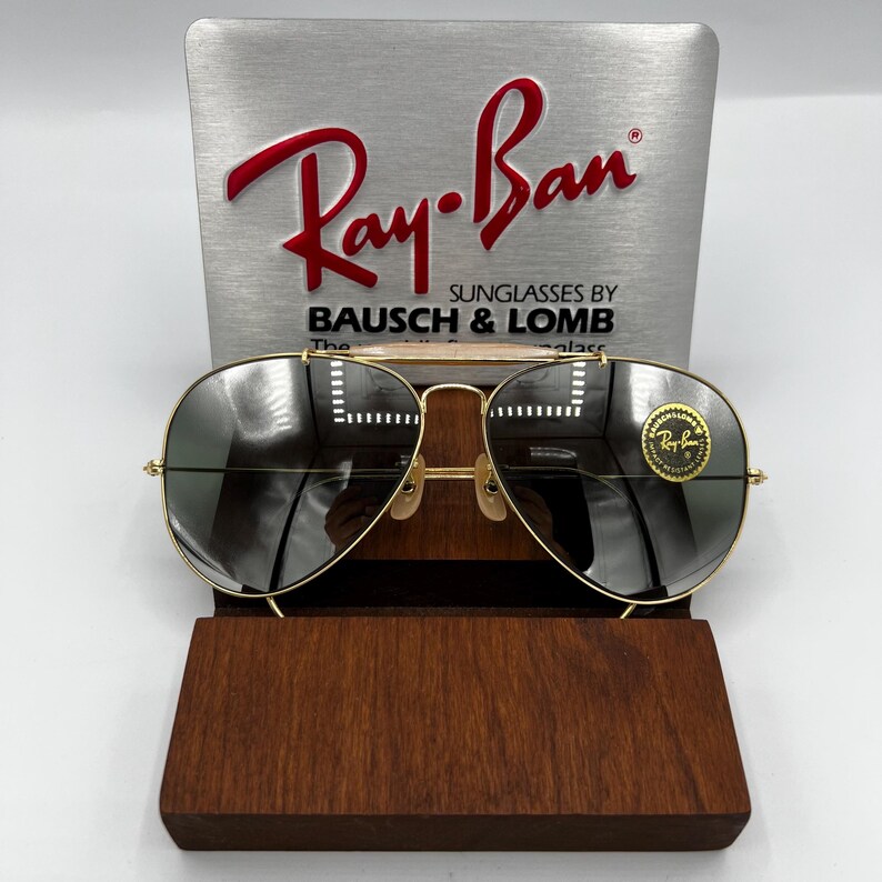 May include: Gold-framed Ray-Ban sunglasses with dark lenses, displayed on a wooden stand. The Ray-Ban logo is visible on the top of the frame and on the lens. The sunglasses are set against a silver background with the Ray-Ban logo and "Sunglasses by Bausch & Lomb".