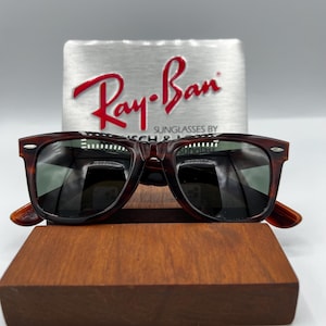 Puede incluir: Gafas de sol con estampado de carey y lentes oscuras, expuestas sobre un bloque de madera. El logotipo de Ray-Ban es visible sobre un fondo plateado. Las gafas tienen una montura rectangular cl&aacute;sica.