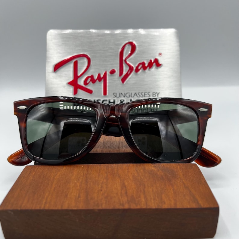 Puede incluir: Gafas de sol con estampado de carey y lentes oscuras, expuestas sobre un bloque de madera. El logotipo de Ray-Ban es visible sobre un fondo plateado. Las gafas tienen una montura rectangular cl&aacute;sica.