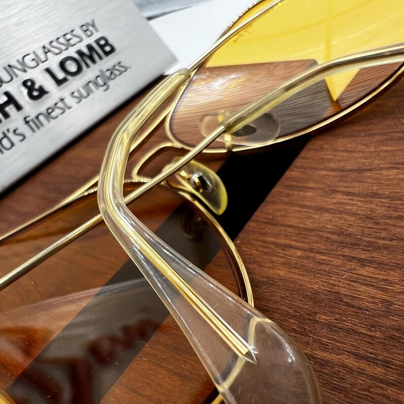 Puede incluir: Gafas de sol de metal en tono dorado con lentes tintadas en amarillo y puntas de patilla de acetato transparente. La montura presenta una construcci&oacute;n de alambre fino. Una placa plateada muestra el texto SUNGLASSES BY BAUSCH & LOMB.