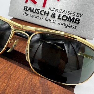 Puede incluir: Primer plano de gafas de sol con montura dorada y lentes oscuras. El texto "SUNGLASSES BY BAUSCH & LOMB The world's finest sunglass." es visible en una placa plateada. Las gafas de sol descansan sobre una superficie de madera.