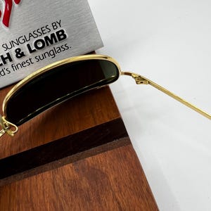 Puede incluir: Gafas de sol con montura dorada y lentes oscuras. Las gafas de sol est&aacute;n sobre una superficie de madera. El texto "SUNGLASSES BY B&LOMB" es visible en el fondo.