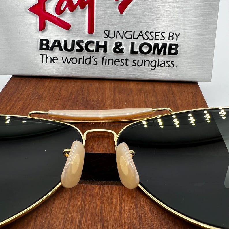 May include: Gold-rimmed sunglasses with black lenses. The sunglasses have a double bridge and light-colored nose pads. The text "Ray-Ban" is in red at the top, and below it, the text reads "SUNGLASSES BY BAUSCH & LOMB The world's finest sunglass."