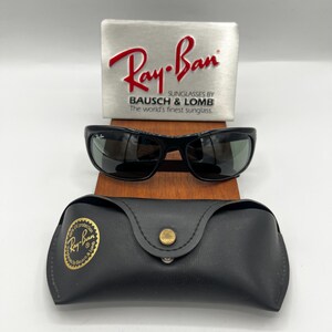 May include: Black Ray-Ban sunglasses with a matching black case and a silver display stand. The Ray-Ban logo is prominently displayed on the stand and case. The sunglasses have a sleek, modern design.