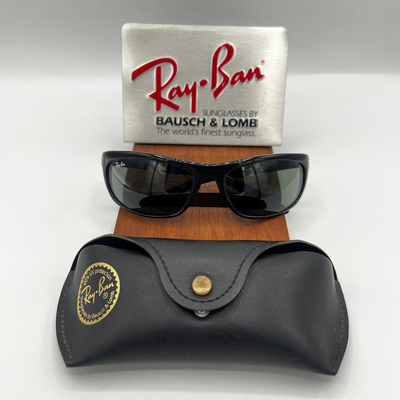 May include: Black Ray-Ban sunglasses with a matching black case and a silver display stand. The Ray-Ban logo is prominently displayed on the stand and case. The sunglasses have a sleek, modern design.