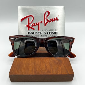 Puede incluir: Gafas de sol marrones carey con lentes oscuras, expuestas sobre un soporte de madera. El logotipo de Ray-Ban es visible sobre un fondo plateado. Las gafas de sol son de estilo cl&aacute;sico, adecuadas para el uso diario.