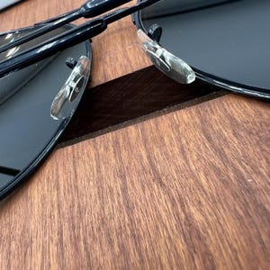 Pu&ograve; includere: Un paio di occhiali da sole neri con un ponte nasale in plastica trasparente e aste in metallo nero. Gli occhiali sono piegati a met&agrave; e sono su una superficie di legno.