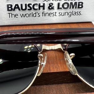 May include: Close-up of Bausch & Lomb sunglasses. The sunglasses have a dark brown frame with gold-colored accents and dark lenses. The text "BAUSCH & LOMB The world's finest sunglass." is visible above the sunglasses.