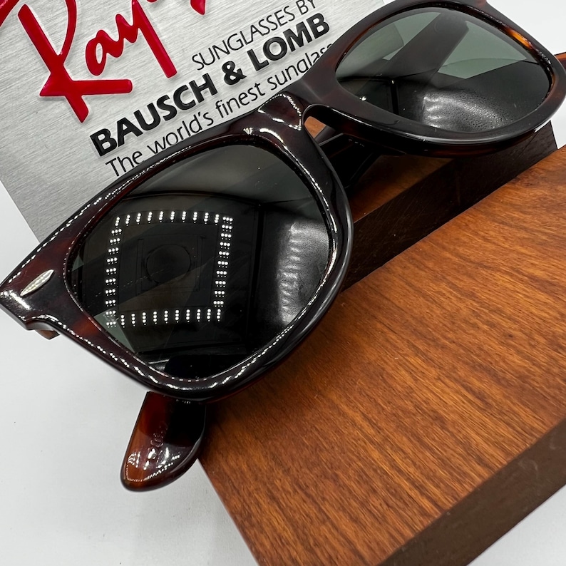 Puede incluir: Gafas de sol carey marr&oacute;n con lentes oscuras, sobre un expositor de madera. El logotipo de Ray-Ban es visible, junto con el texto "SUNGLASSES BY BAUSCH & LOMB The world's finest sunglasses."