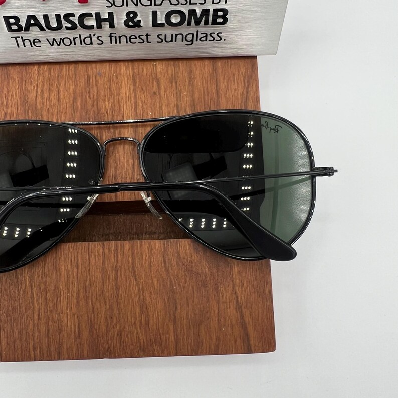 Pu&ograve; includere: Occhiali da sole aviator neri con lenti verdi. Gli occhiali sono su un espositore in legno. Il nome del marchio "Ray-Ban" &egrave; visibile sulla lente destra.