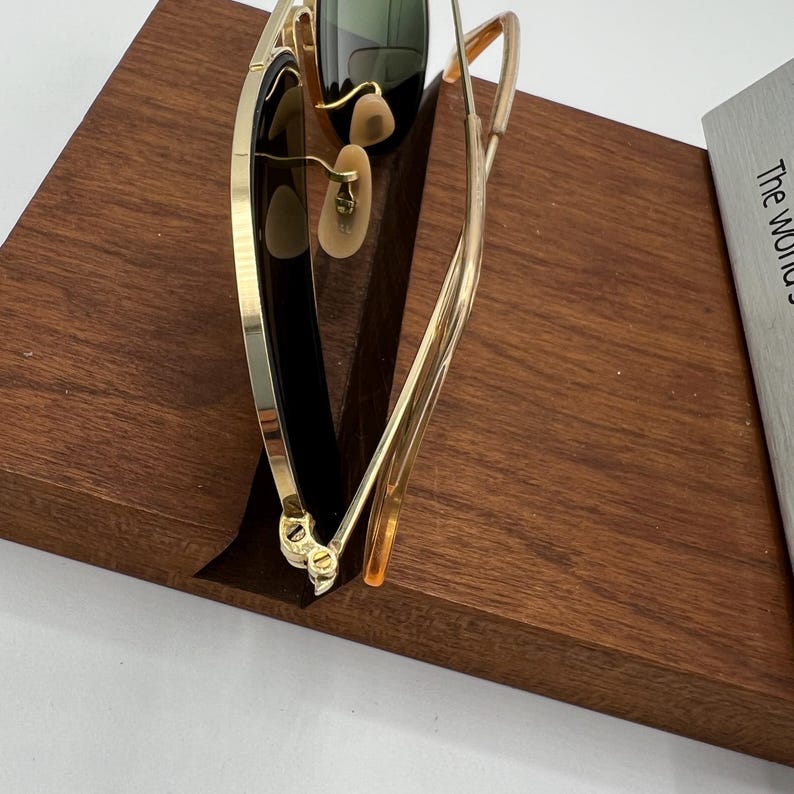 K&ouml;nnte beinhalten: Goldfarbene Aviator-Sonnenbrille mit dunkelgr&uuml;nen Gl&auml;sern und einem braunen Holzst&auml;nder.