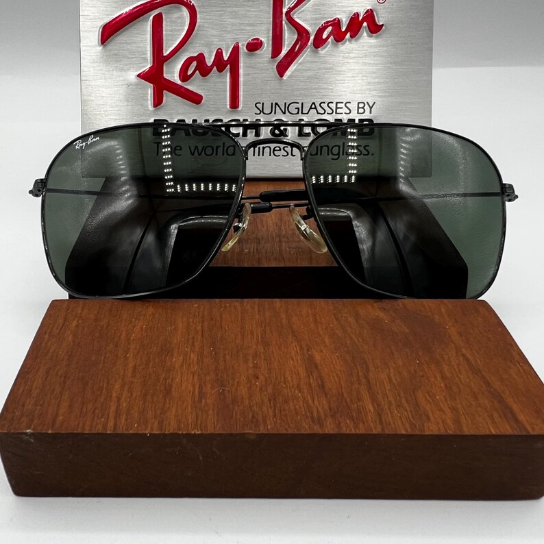 K&ouml;nnte beinhalten: Schwarze Ray-Ban Sonnenbrille mit gr&uuml;nen Gl&auml;sern liegt auf einem Holzdisplay. Die Sonnenbrille hat eine klassische Aviator-Form mit einem d&uuml;nnen schwarzen Rahmen. Das Ray-Ban-Logo ist oben auf dem Rahmen sichtbar. Der Hintergrund zeigt das Ray-Ban-Logo und Text.
