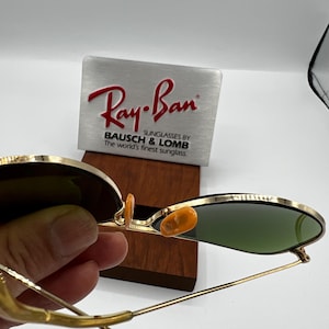 May include: Gold-framed Ray-Ban sunglasses with green lenses. The sunglasses are held in front of a silver and wooden display with the Ray-Ban logo and text: "SUNGLASSES BY BAUSCH & LOMB The world's finest sunglass."