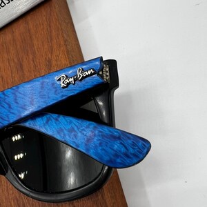 Op de afbeelding: Zwarte zonnebril met blauw patroon op de pootjes. Het Ray-Ban logo is zichtbaar op de blauwe poot. De zonnebril heeft een klassiek ontwerp en is geschikt voor buitenactiviteiten.