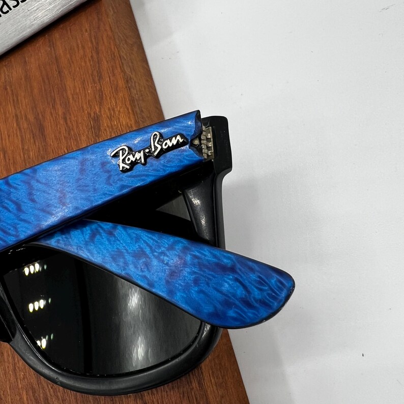 Op de afbeelding: Zwarte zonnebril met blauw patroon op de pootjes. Het Ray-Ban logo is zichtbaar op de blauwe poot. De zonnebril heeft een klassiek ontwerp en is geschikt voor buitenactiviteiten.