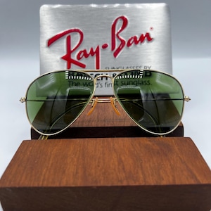 May include: Gold-rimmed aviator sunglasses with green lenses. The sunglasses are displayed on a wooden stand, with a silver background featuring the Ray-Ban logo in red. The text on the background reads "The world's finest sunglass."