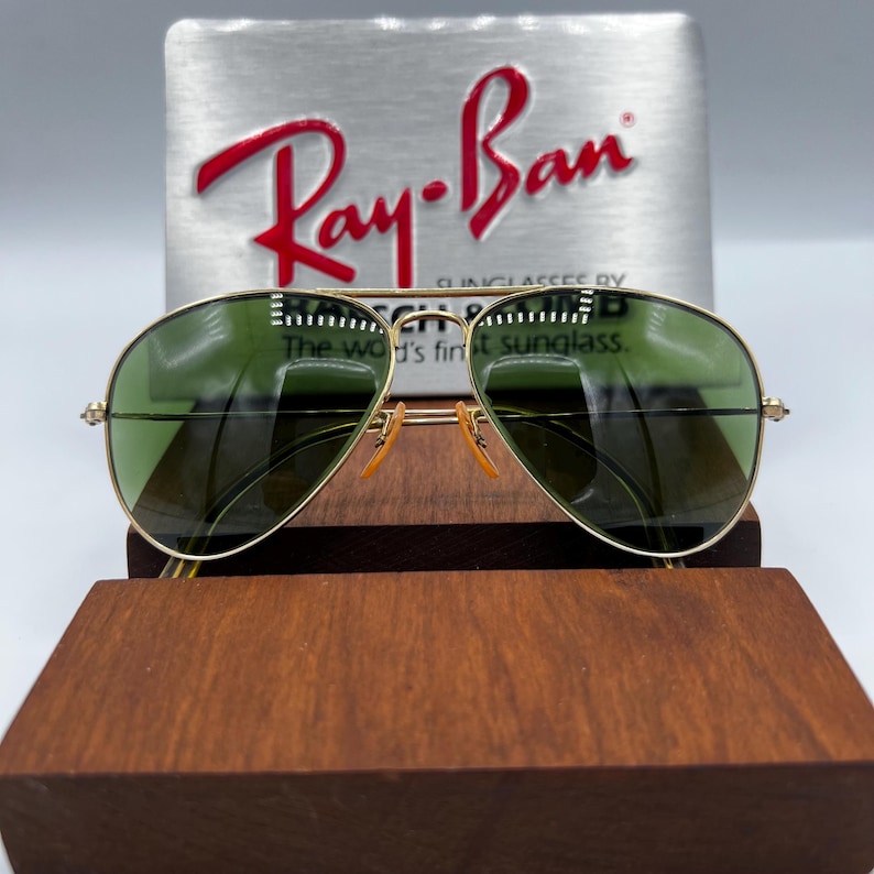 May include: Gold-rimmed aviator sunglasses with green lenses. The sunglasses are displayed on a wooden stand, with a silver background featuring the Ray-Ban logo in red. The text on the background reads "The world's finest sunglass."