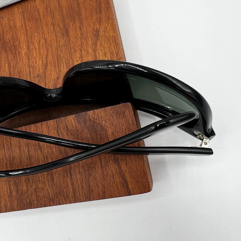 May include: Black sunglasses with dark lenses. The sunglasses are resting on a wooden surface. The frames are thick and have a glossy finish. The arms of the sunglasses are extended.