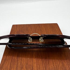 May include: A pair of eyeglasses with a dark burgundy frame and gold-colored nose bridge. The glasses are resting on a wooden surface, with the lenses facing upwards. The overall design is sleek and modern.