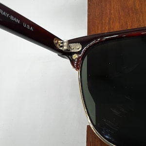 May include: Close-up of Ray-Ban sunglasses with a dark brown frame and black lenses. The hinge is silver, and the arm is marked "RAY-BAN U.S.A." The sunglasses have a classic design.