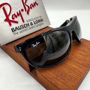 May include: Black Ray-Ban sunglasses with dark tinted lenses. The sunglasses are displayed on a wooden stand, with the Ray-Ban logo visible on the frame. The box behind the sunglasses reads "Ray-Ban" and "SUNGLASSES BY BAUSCH & LOMB".