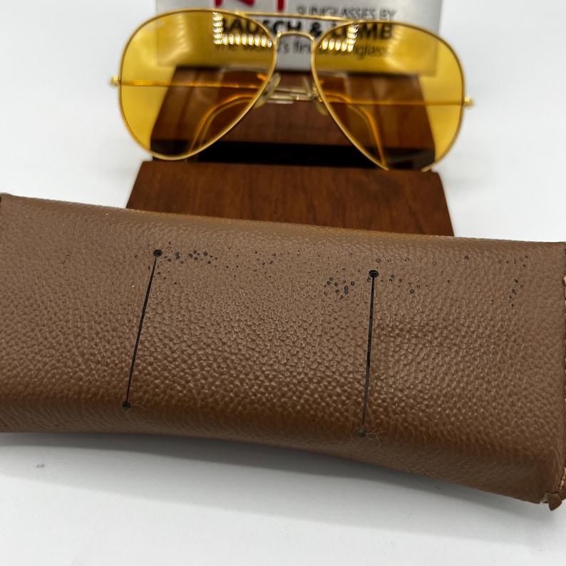 Puede incluir: Gafas de sol tipo aviador con montura de metal dorado y lentes tintadas en amarillo, situadas detr&aacute;s de un estuche de cuero marr&oacute;n texturizado. Las gafas presentan una barra superior con el texto Bausch & Lomb, the world's finest sunglasses.