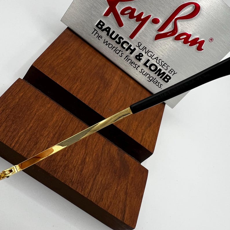 Puede incluir: Primer plano de gafas de sol Ray-Ban con montura dorada y patillas negras, apoyadas en un expositor de madera. El logotipo de Ray-Ban y el texto "BAUSCH & LOMB" son visibles en una etiqueta plateada.