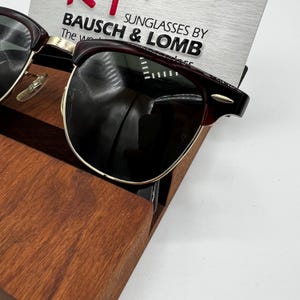 May include: Sunglasses by Bausch & Lomb with a dark burgundy frame and gold accents. The lenses are dark, and the sunglasses are displayed on a wooden stand. The text "SUNGLASSES BY BAUSCH & LOMB" is visible.