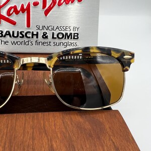 May include: Tortoiseshell-patterned sunglasses with gold-colored frames and brown lenses. The sunglasses are displayed in front of a sign that reads "Ray-Ban" and "Sunglasses by Bausch & Lomb, The world's finest sunglass."