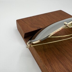 K&ouml;nnte beinhalten: Eine silberfarbene Sonnenbrille mit goldenen Akzenten, zusammengeklappt und auf einer Holzoberfl&auml;che liegend.